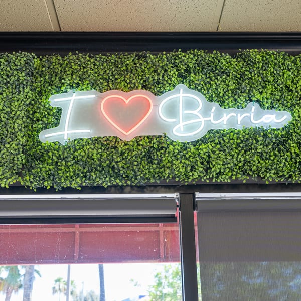 Neon sign spelling "I heart Birria" on a green faux plant wall above a window.