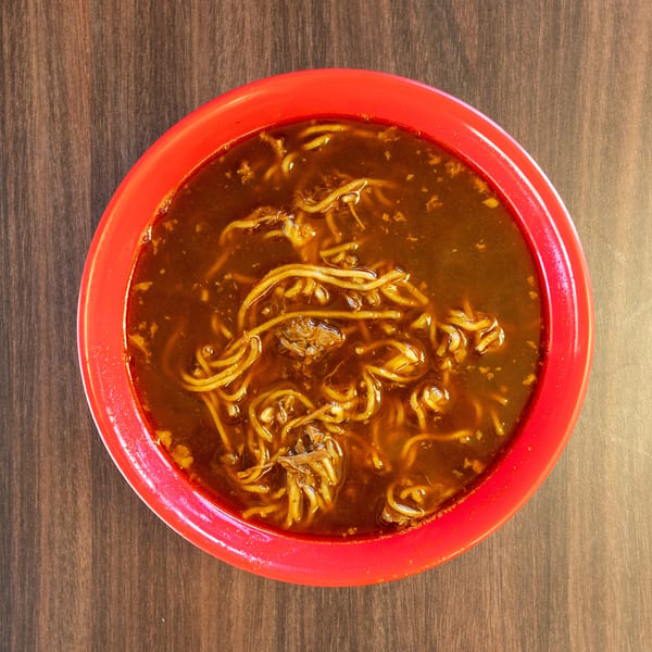 Overhead view of a red bowl filled with a dark broth and noodles on a wooden table.