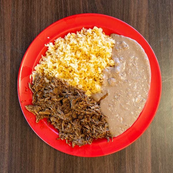 A red plate holds shredded beef, yellow rice, and refried beans on a wooden table.