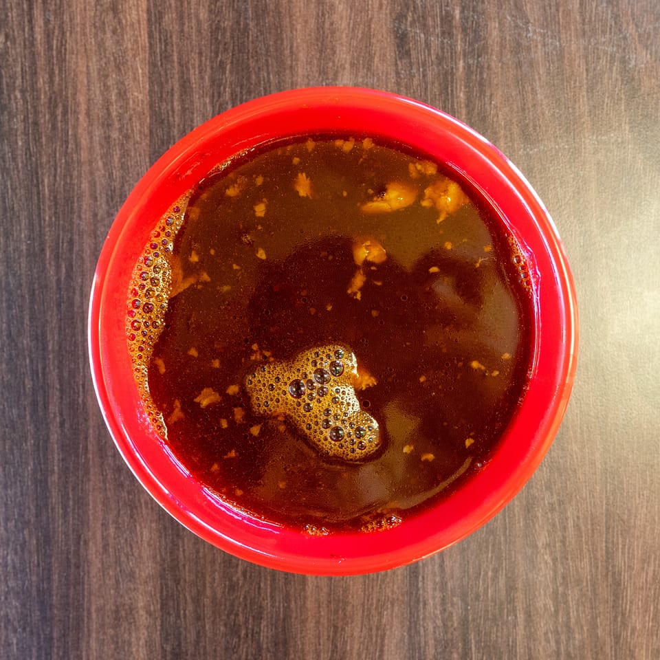 Overhead view of a red bowl filled with dark brown soup, showing small bubbles and food bits.