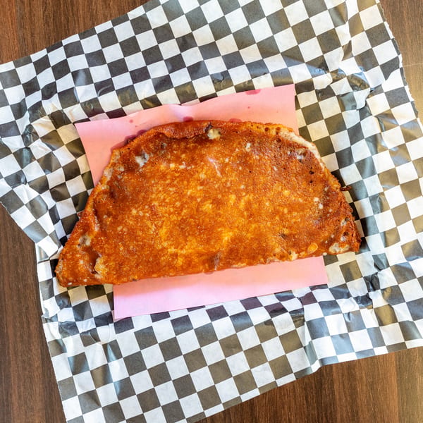 Crispy, golden-brown quesadilla on pink paper and checkered wax paper, on a dark wooden table.