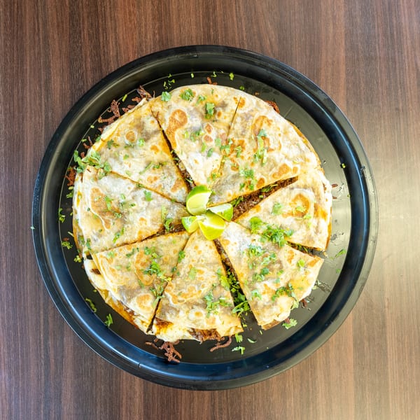 Overhead view of a large quesadilla cut into wedges, garnished with cilantro and lime, on a black platter.