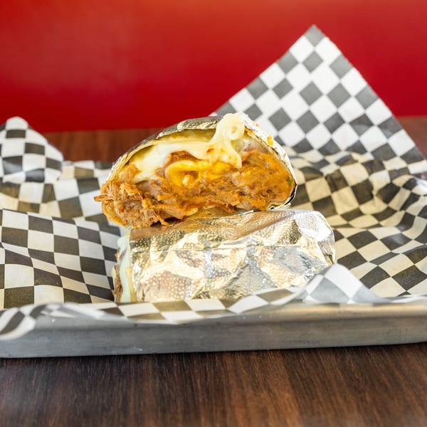 A foil-wrapped burrito, cut in half, revealing melted cheese and savory filling on checkered paper.