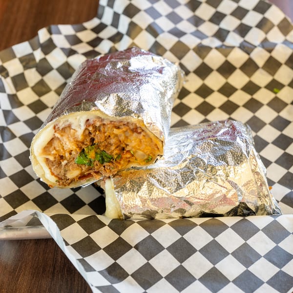 Two foil-wrapped burritos, one cut open to show meat, rice, and cheese, in a checkered basket.