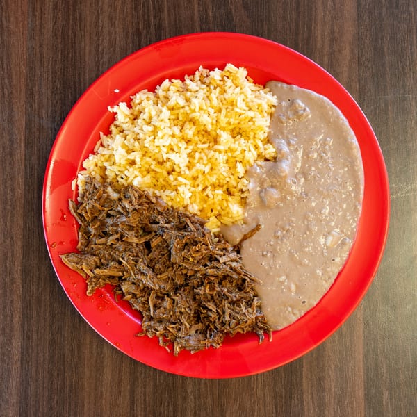 Red plate with shredded beef, yellow rice, and refried beans on a wooden table.